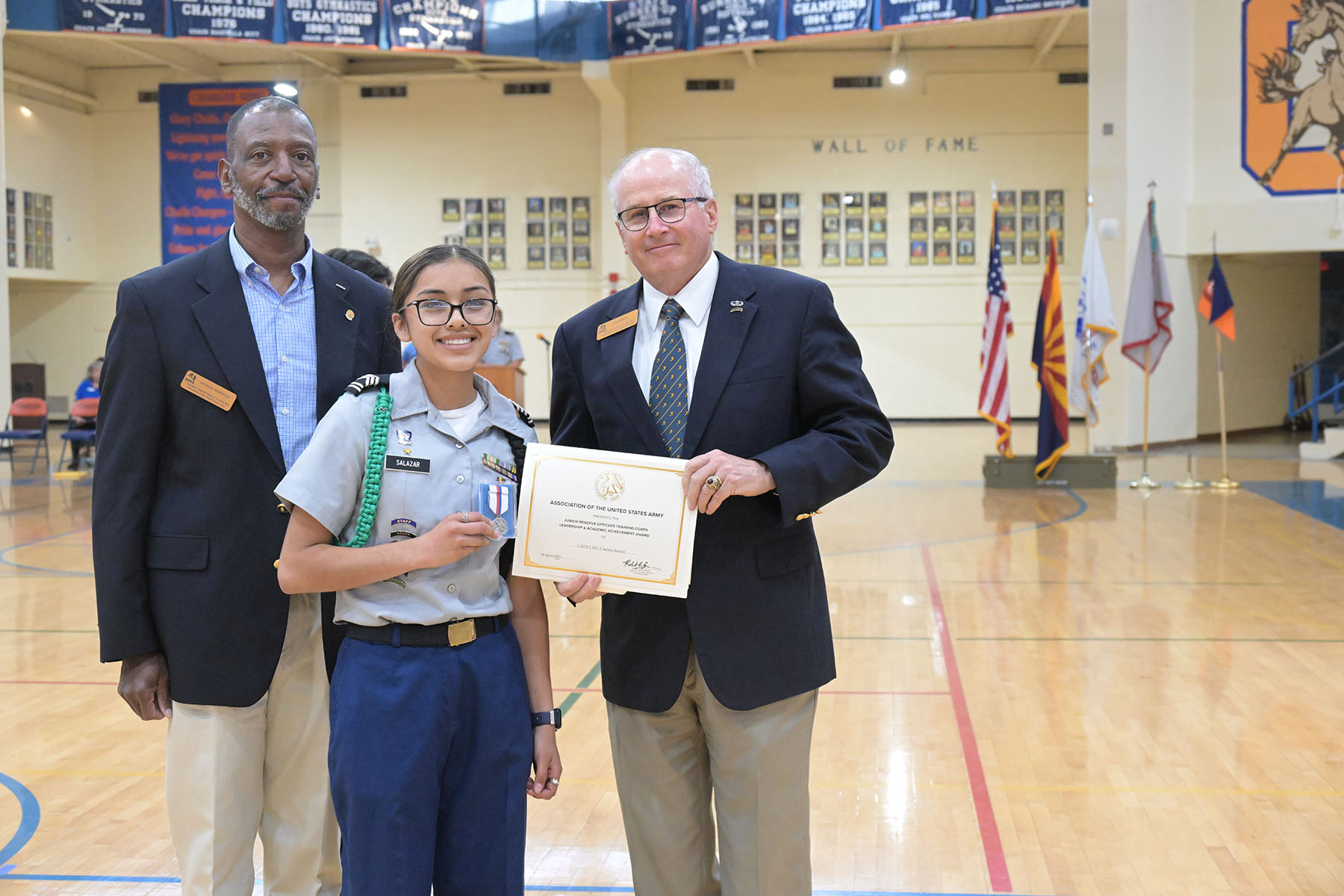 A teen girl in glasses stands between two men, showing off her certificate