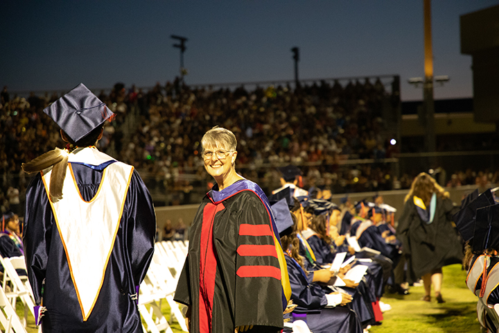 A woman in graduation regalia smiles as she walks up the aisle past a graduate