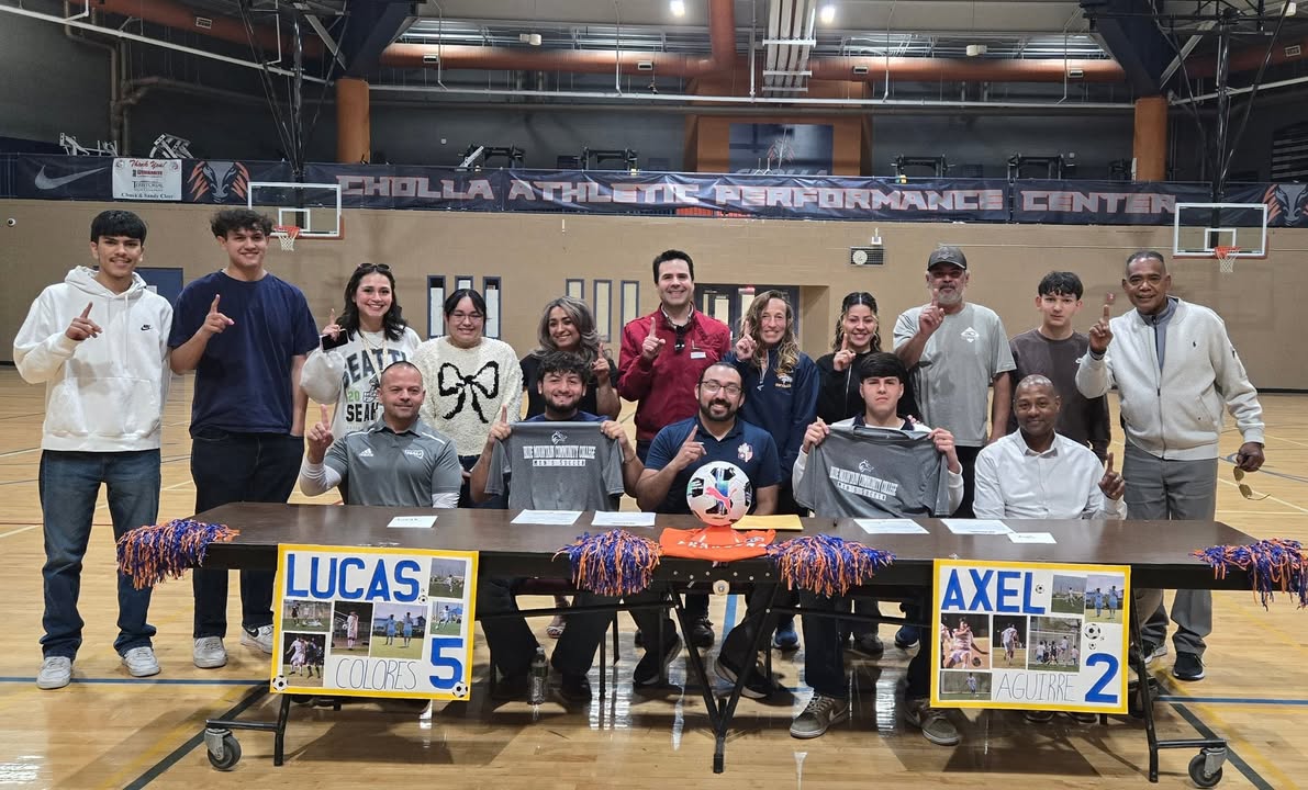 Two students hold up their college shirts while coaches, teachers and family members stand around them
