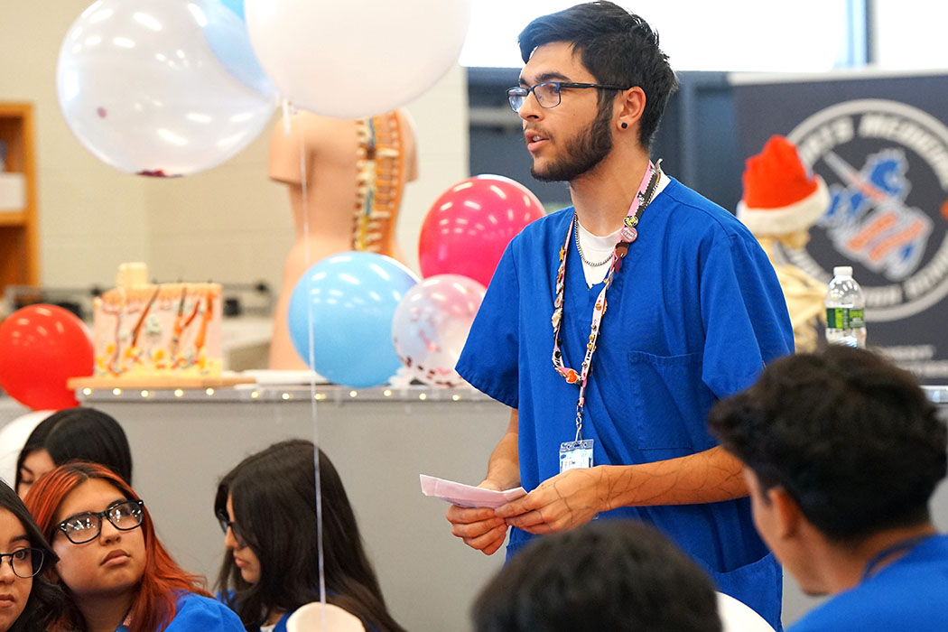 A boy in blue scrubs and glasses stands up to address his classmates