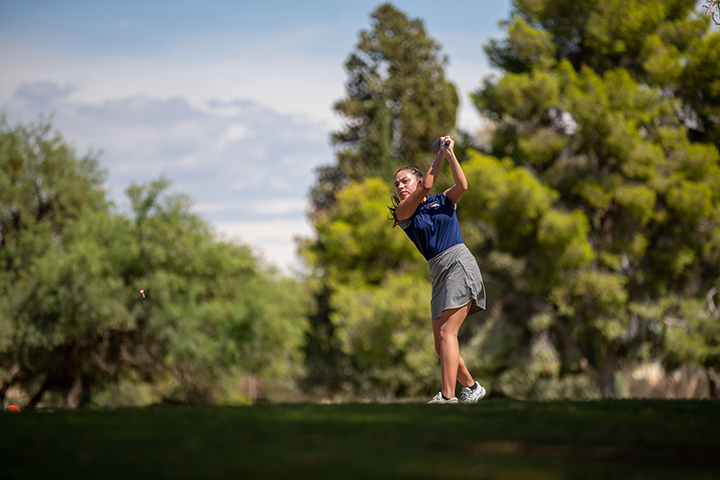 A girl in a navy polo and gray skort swings a golf club on the green
