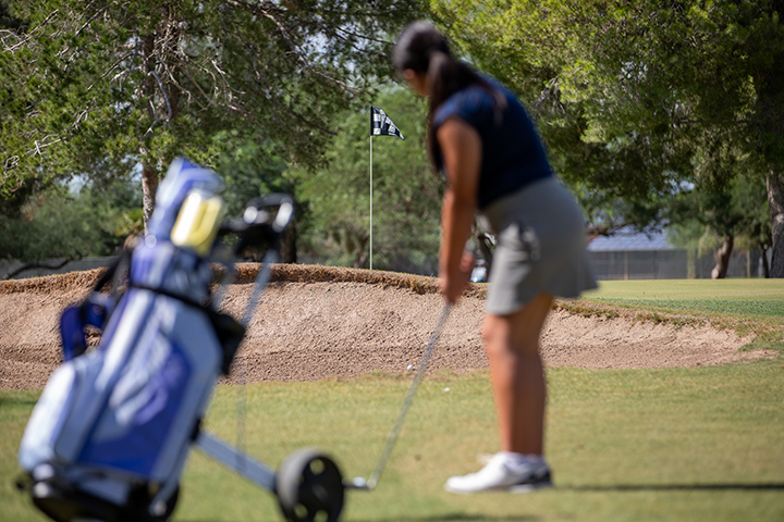A girl in a navy polo and gray skort concentrates on hitting her golf ball into the hole