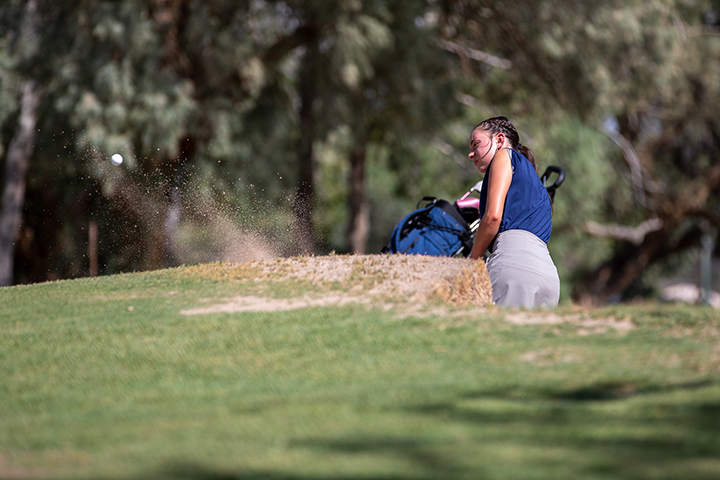 A girl in a navy tank and gray skirt swings her golf club in the sand trap