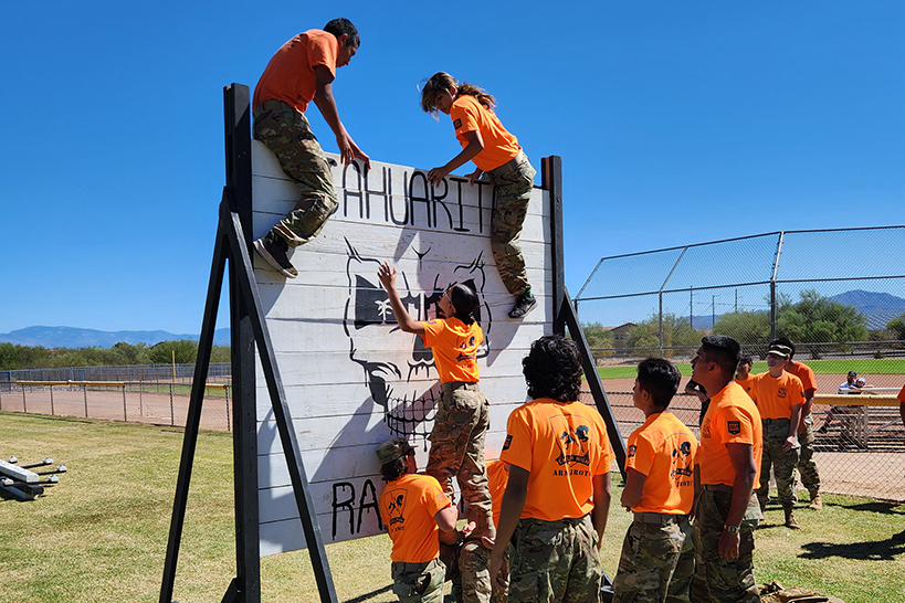 JROTC students in orange shirts climb over a wooden wall