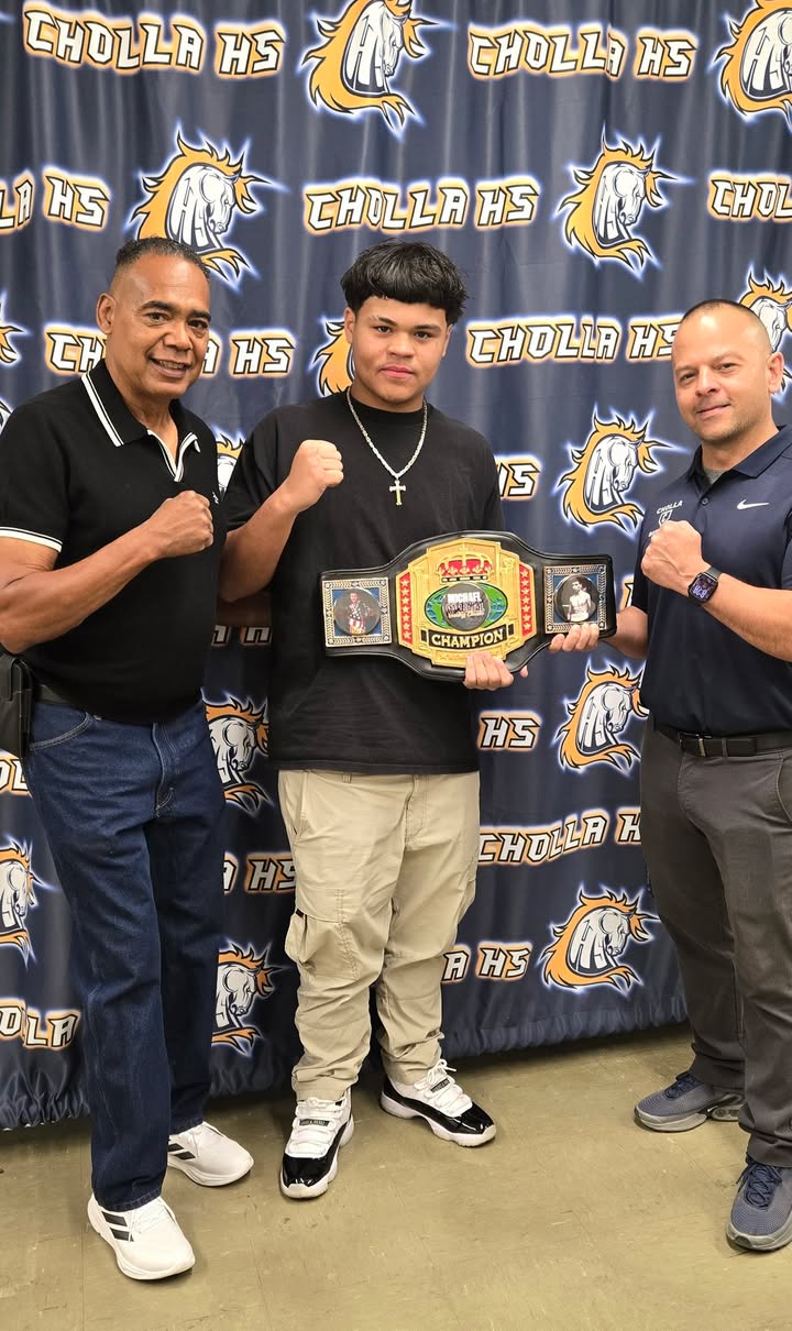 A teen boy holds up his boxing championship belt, surrounded by his two coaches
