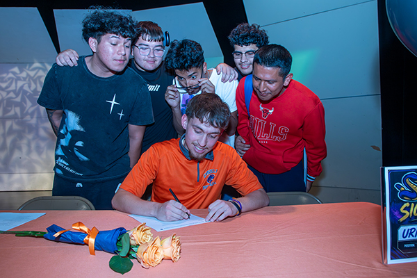 A teen boy smiles as he signs his college letter of intent, as friends look over his shoulder