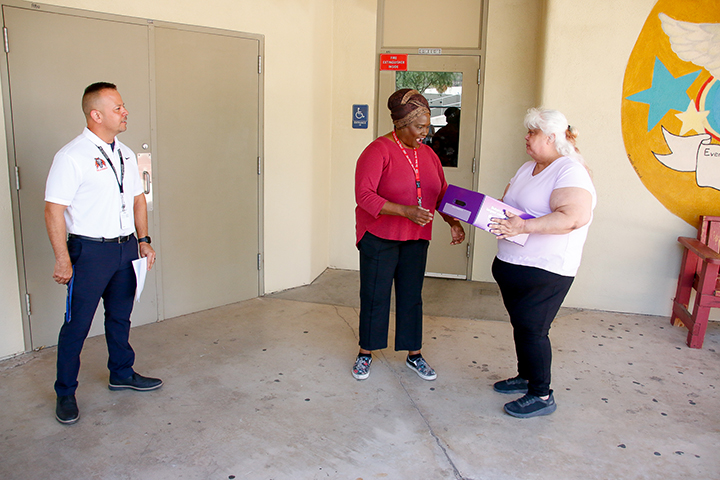 A woman wearing red, center, gasps in surprise as a woman, right, hands her a purple box. A man, left, observes.