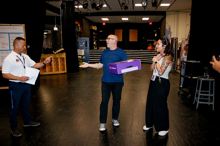 A man, left, points to a man, center, holding a purple box. A woman, right, holds her arms to her chest in awe