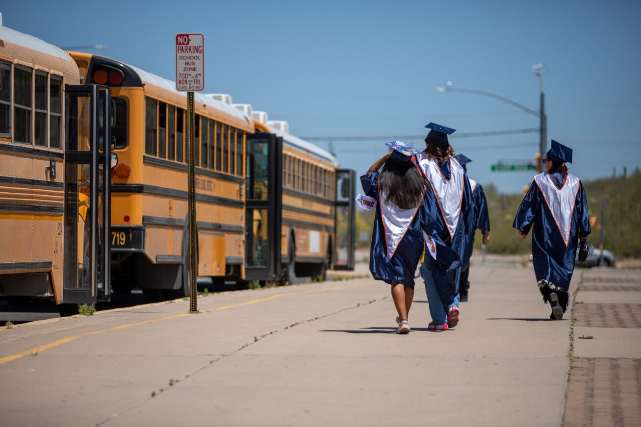 Cholla grads walk in their caps and gowns towards a line of waiting school buses