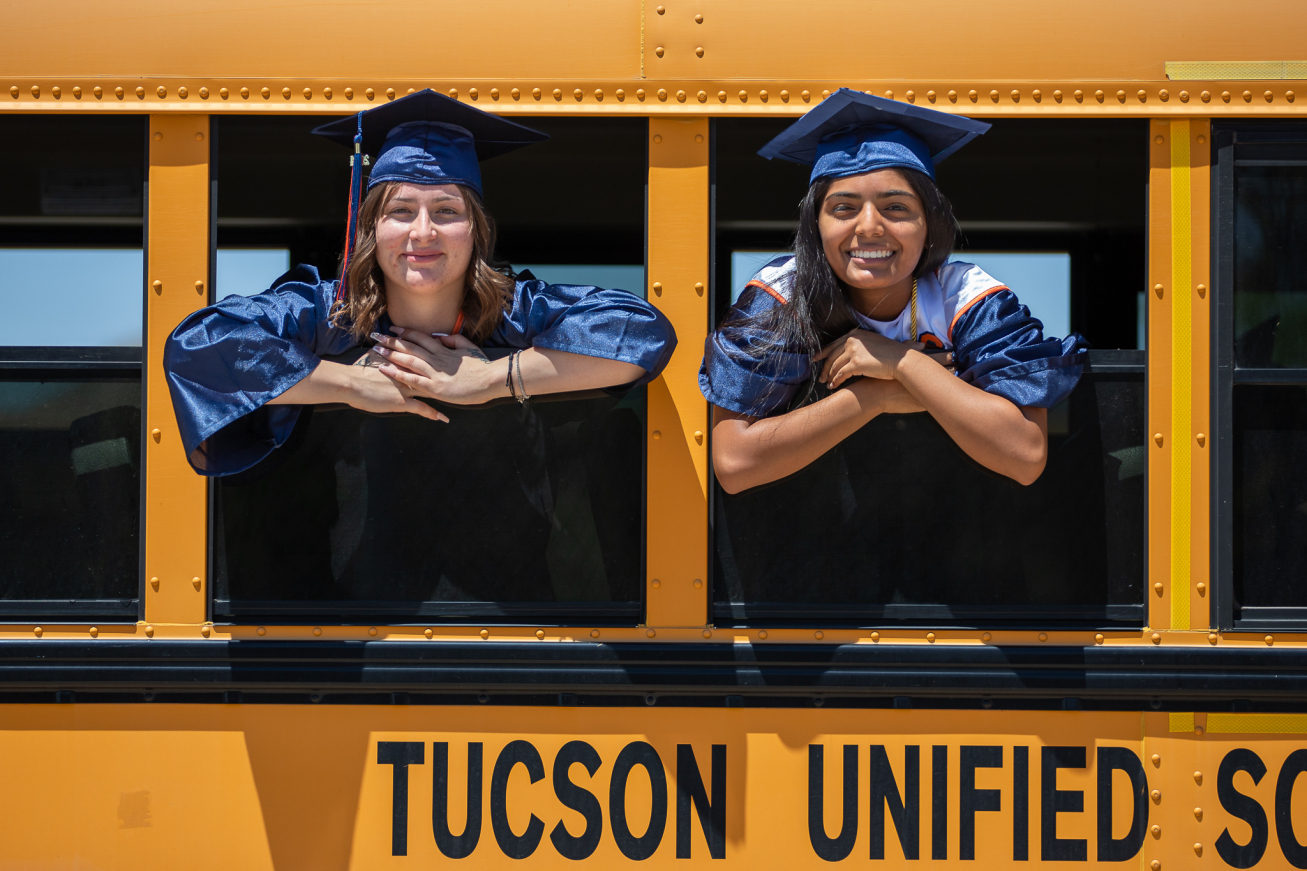 Two grads smile in their caps and gowns as they hang out the windows of a school bus