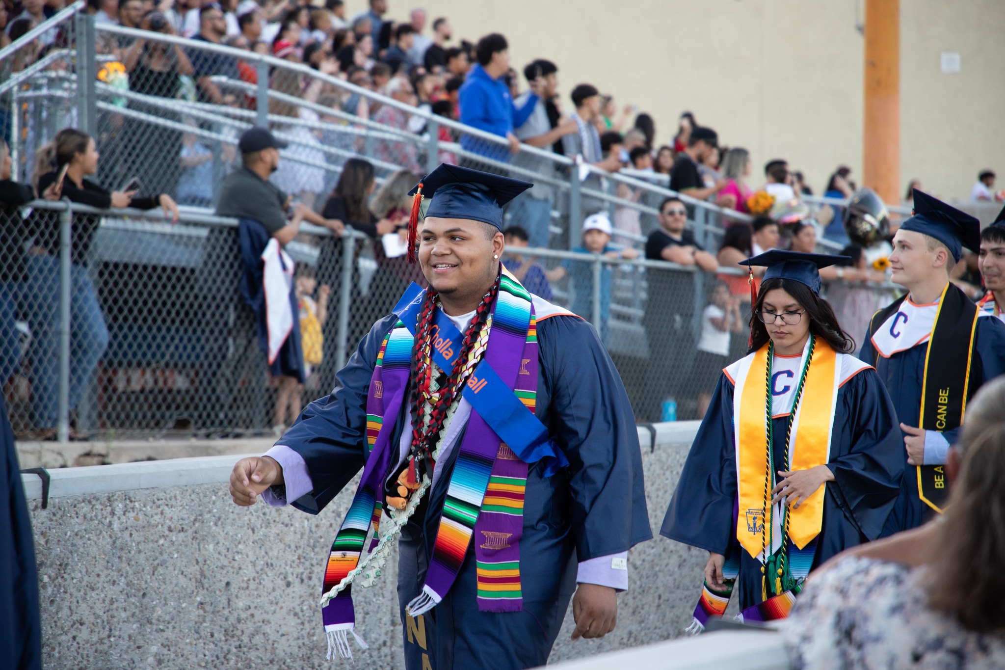 A Cholla grad smiles in his cap and gown as he makes his way onto the football field